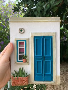 a hand holding up a miniature house with blue shutters and potted plants in front of it