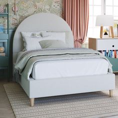 a white bed sitting next to a window with curtains on the windowsill and bookshelves in front of it