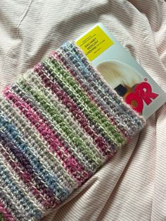 a crocheted book cover laying on top of a white bed with pink, blue, and green stripes