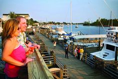 two people standing on a dock with boats in the water and one person holding a drink