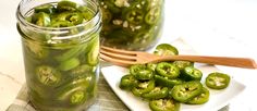 pickled green peppers sit on a white plate next to a glass jar and wooden spoon