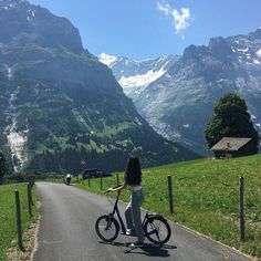 a woman is riding her bike down the road in front of some mountains and green grass