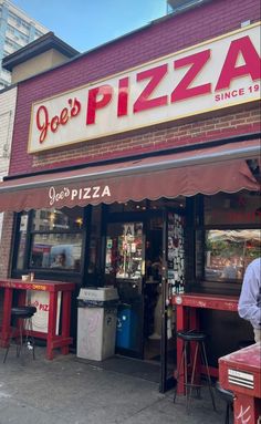 a man standing in front of a pizza shop next to a red table and chairs