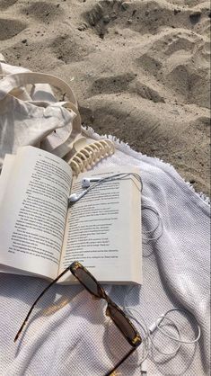 an open book sitting on top of a white blanket next to sunglasses and headphones