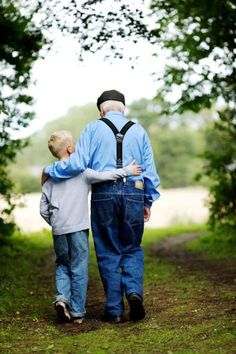 an older man and young boy walking down a dirt path in the woods with their backs to each other