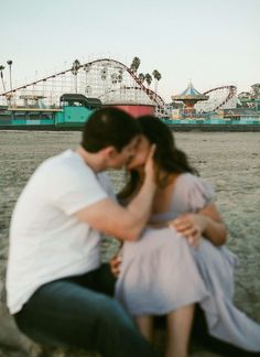 a man kneeling down next to a woman on top of a sandy beach near roller coasters