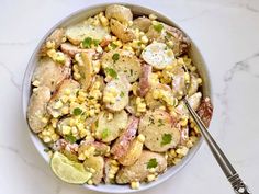 a bowl filled with corn and potatoes on top of a white table next to a fork