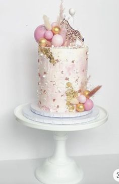 a pink and gold birthday cake on a white pedestal with some balloons in the background