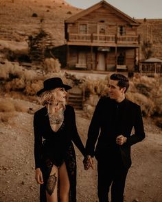 a man and woman holding hands while walking down a dirt road in front of a house