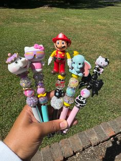 a hand holding several toy animals and pens in front of a brick wall on the grass