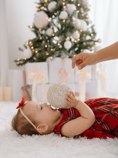 a baby is laying on the floor near a christmas tree and holding a silver ornament