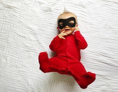 a baby wearing a mask laying on top of a white sheet covered bed with his hands in his mouth