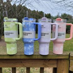 four different colored coffee mugs sitting on top of a wooden fence rail in front of trees