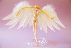 a doll with white wings is standing on a glass base in front of a pink background