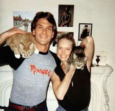 a man and woman holding two cats in front of a fireplace with pictures on the wall behind them