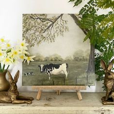 a painting of a cow and two rabbits on a shelf next to a potted plant