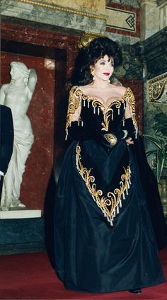 a woman in a black and gold dress standing next to a statue wearing a tiara