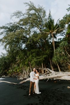 a man and woman standing in front of trees on the black sand beach with their arms around each other