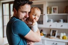 a man holding a baby in his arms and looking at the camera while standing in a kitchen