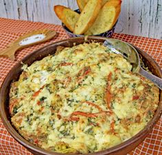 a casserole dish with cheese and vegetables in it on a table next to slices of bread