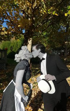 a man and woman dressed up in costumes kissing under a tree with leaves on the ground