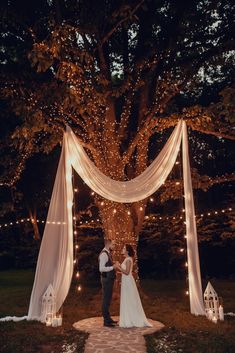 a bride and groom standing in front of a tree with fairy lights on the branches