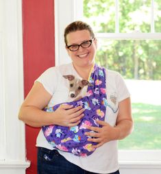 a woman is holding a small dog in her arms while wearing a purple floral sling