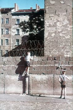 two children standing on the edge of a wall with barbed wire around them and buildings in the background