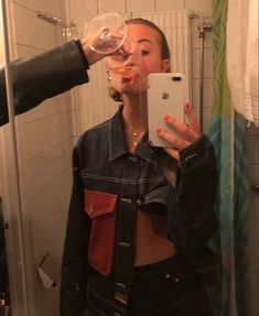 a woman drinking from a wine glass while standing in front of a mirror with her phone