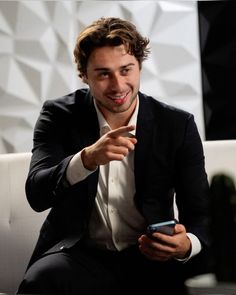 a man in a black suit and white shirt is holding a smart phone while sitting on a couch