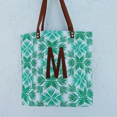 a green and white tote bag hanging from a hook on a wall with palm leaves