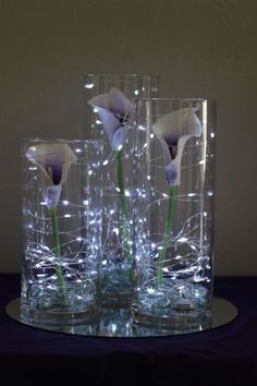 three glass vases sitting on top of a table with flowers in them and lights behind them