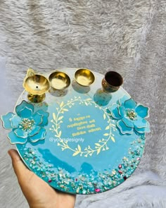 a hand holding a blue plate with three gold cups on it and two spoons in the middle