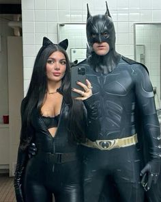 two people dressed up as batman and catwoman standing in a public restroom, one holding a cell phone