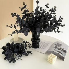 a vase filled with black flowers next to an open book on a white table top