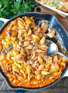 a skillet filled with pasta and meat on top of a wooden table next to bread