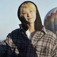 a young man with blonde hair wearing a black and white plaid shirt standing in front of a hot air balloon