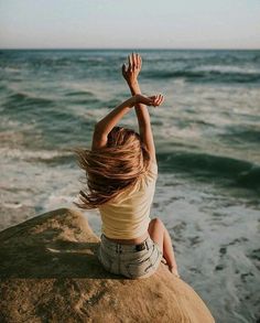 a woman sitting on top of a rock next to the ocean with her arms in the air