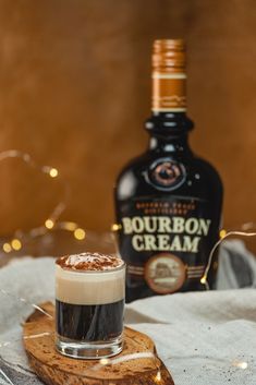 a bottle of bourbon cream sitting next to a small cup on a wooden board with lights in the background