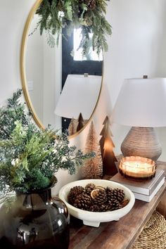 a table topped with a bowl filled with pine cones next to a mirror and lamp