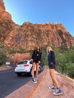 two women standing on the side of a road next to a white car and mountains