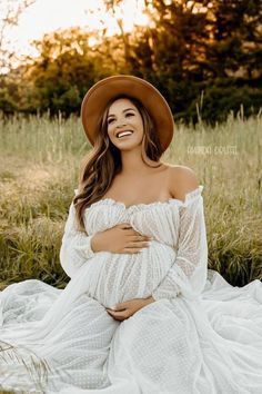 a pregnant woman wearing a white dress and brown hat smiles while sitting in the grass