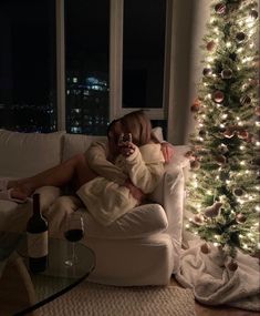 a woman sitting on a couch holding a wine glass in front of a christmas tree