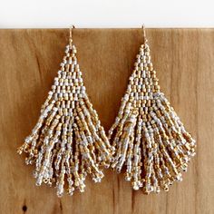 Sokko Hasimbish in the Choctaw language translates to thick tail. These hand-beaded earrings with a structured brick stitch diamond-shaped top have twenty-four double-layered fanned fringe that ends in an ornate diamond shape. Sokko Hasimbish contains approximately 564 delicate glass beads in each earring. white matte galvanized gold, opal white, 24K gold-filled ear wires mineral sterling silver, metallic gold, silver-lined crystal, gold-lined crystal, 24K gold-filled ear wires spruce matte galv Artisan Gold Jewelry With Fringe, Silver Handwoven Dangle Jewelry, Choctaw Language, Opal White, Silver Line, Gold Line, Gold Orange, Brick Stitch, Diamond Shaped