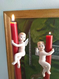 two candles with angel figurines on them sitting in front of a framed painting