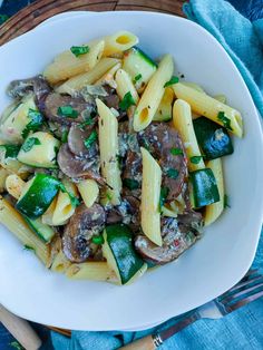 pasta with mushrooms and zucchini in a white bowl on a blue cloth next to silverware