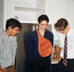 three young men standing in a kitchen looking at something on the refrigerator freezer door