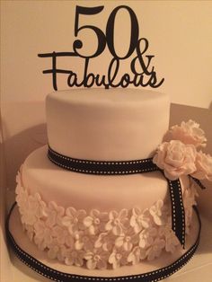 a white cake with black lettering and flowers on the bottom tier is sitting in front of a sign that reads 50 fabulouss