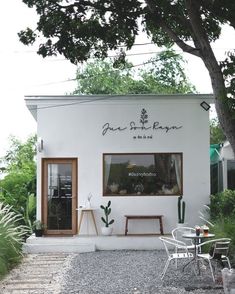 the outside of a small white building with tables and chairs around it on graveled area next to trees