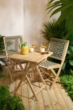 two chairs and a table on a wooden deck in front of some plants, with drinks